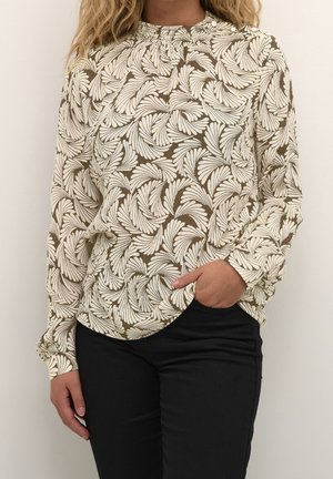 Kobieta ubrana w bluzkę z długim rękawem z beżowo-brązowymi abstrakcyjnymi wzorami liści oraz w czarne spodnie, z ręką częściowo w kieszeni.