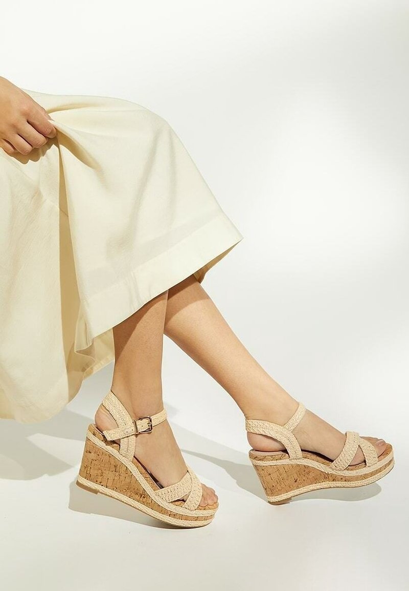 Personne portant des sandales compensées en tissu beige avec brides à la cheville et une jupe crème claire, assise devant un fond blanc uni.