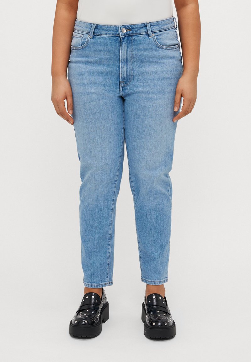 Hellblaue Denim-Jeans mit hoher Taille, geradem Schnitt, subtiler Ausbleichung und Fünf-Taschen-Design, kombiniert mit schwarzen, mit Nieten verzierten Slippern.