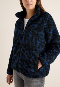 Giacca in pile a fantasia nero e blu con colletto alto, chiusura con zip e tasche frontali, realizzata in un tessuto morbido e texturizzato.