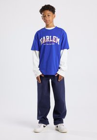 Blue cotton t-shirt with “HARLEM” printed in white and red, layered over a long-sleeve white shirt. Worn with dark navy pants and white sneakers.