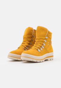 Bottes cheville en daim jaune avec un intérieur en peluche, dotées d'œillets métalliques, de lacets épais et d'une semelle blanche épaisse avec des accents jaunes.