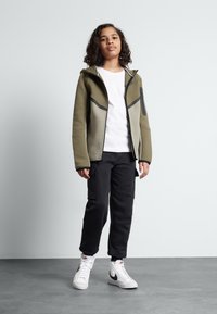 Felpa con zip color verde oliva con dettagli neri, pannelli grigi e tasche frontali, abbinata a pantaloni della tuta neri e sneakers bianche alte.