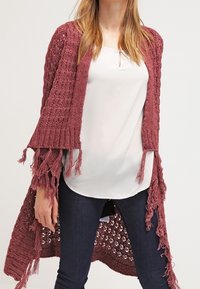 Cardigan en tricot bordeaux avec une ouverture sur le devant, des accents à franges et un design loose et texturé. Associé à un haut blanc et un jean bleu.