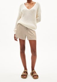 Pull en tricot beige clair avec col en V et manches amples, associé à un short beige et des sandales marron à enfiler. Texture lisse dans l'ensemble.