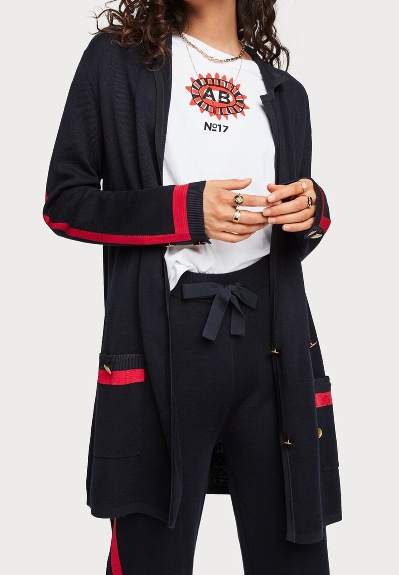 Granatowy kardigan z czerwonymi paskami, otwarty z przodu, z naszywanymi kieszeniami; miękki dzianinowy materiał, złote akcenty, noszony na białej koszulce z nadrukiem i spodniach.