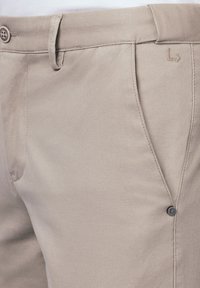 Beżowe spodnie wykonane z teksturowanej tkaniny, z zapięciem na guzik i subtelnym detalem logo w pobliżu pasa. Mają klasyczny krój.
