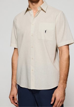 Man draagt een beige overhemd met korte mouwen en een borstzak met een klein zwart golferlogo, gecombineerd met een donkere broek.