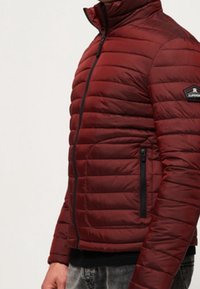 Červená prošívaná bunda s vysokým límcem, vertikálním prošíváním, černými kapsami na zip a logem na horní části rukávu.