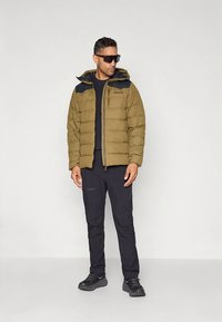 Olivgrön vadderad jacka med svarta detaljer och huva, tillsammans med svarta långa byxor och mörka sneakers. Mjuk textur, casual passform.