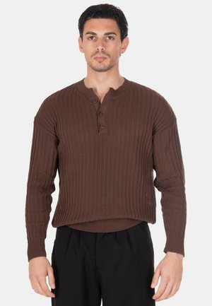 Uomo che indossa un maglione marrone a coste con un patta con tre bottoni, abbinato a pantaloni neri, in piedi davanti a uno sfondo semplice.