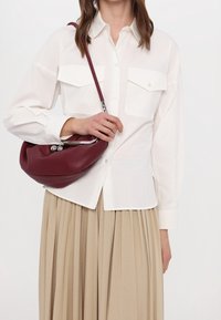 Borsa a tracolla in pelle color bordeaux con forma curva, accessori in metallo argento, abbinata a una camicia bianca con bottoni e una gonna plissettata beige.