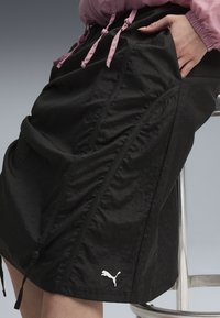 Jupe noire faite en tissu texturé avec des détails froncés, dotée de poches latérales et d'un logo blanc au bas.