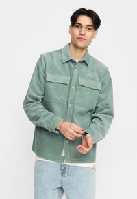 Groene corduroy shirt met een knoopsluiting aan de voorkant, twee borstzakken en lange mouwen, gedragen over een lichtgekleurde t-shirt en jeans.