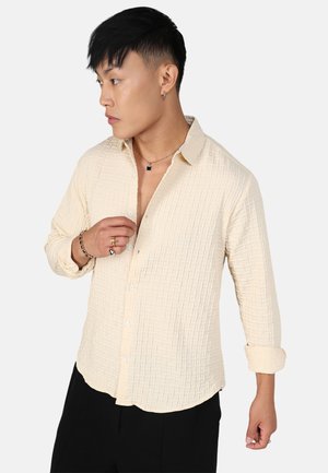 Camisa de botones beige claro con un patrón texturizado, de mangas largas y con un dobladillo redondeado, usada parcialmente desabotonada, sobre un fondo neutro.