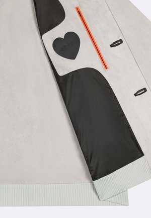 Manica di giacca grigio chiaro con fodera interna nera, tasca con cerniera arancione, toppa a forma di cuore nera e dettaglio polsino a costine.