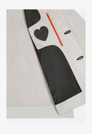 Manica di giacca grigio chiaro con fodera interna nera, tasca con cerniera arancione, toppa a forma di cuore nera e dettaglio polsino a costine.