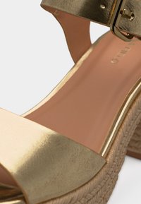 Sandale compensée dorée avec semelle en jute tressé et bride à la cheville ornée d'une boucle dorée, détail en gros plan des parties avant et latérale.