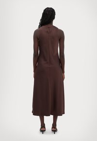 Braunes ärmelloses Midikleid mit glatter Textur, lockerem Schnitt und subtilen Rückennahtdetails. Getragen mit schwarzen Sandalen mit Riemen.