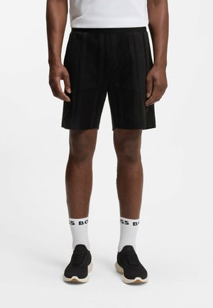 Mężczyzna ubrany w czarne sportowe szorty, białe skarpetki z czarnym napisem, czarne tenisówki i białą koszulkę na tle jednolitego białego tła.