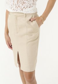 Falda lápiz beige en un tejido texturizado, con una abertura frontal y bolsillos laterales, con un acabado suave y un corte ajustado. Pulsera dorada visible.