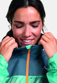 Windbreaker-Jacke in Teal und Grün, mit einem strukturierten Design, orangefarbenem Reißverschluss und hohem Kragen. Am Hals für mehr Komfort angepasst.