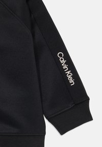 Čierna mikina s rebrovaným manžetovým rukávom, na ktorom je blízko zápästia vytlačené biele logo "Calvin Klein".