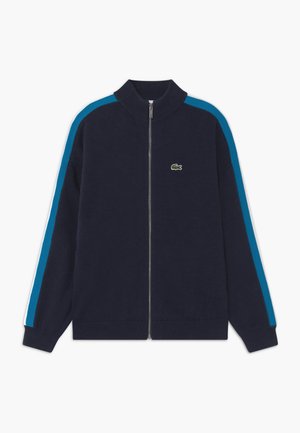 Laivastonsininen vetoketjullinen takki, jossa on resoritettu kaulus, hihansuut ja helma, vaaleansiniset raidat hihoissa ja pieni vihreä krokotiililogo rinnassa.