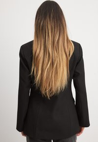 Melna žakete ar pielāgotu dizainu, izturīgām plecu daļām un gludu tekstūru. Garie, taisnie mati daļēji redzami, skats no aizmugures.