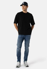 Camisa de algodón negra con bolsillo en el pecho, combinada con jeans de mezclilla azul claro desgastados. Acompañada de una gorra azul marino y zapatillas de correr grises.