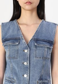 Jeansvest med V-hals, korte ermer og to frontlommer. Har fem hvite knapper og en lys blå vask med synlige sømmer.