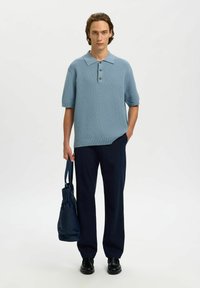 Jeune homme portant un polo en maille bleu clair à manches courtes, un pantalon bleu marine, des chaussures noires, tenant un sac fourre-tout bleu marine dans une main.