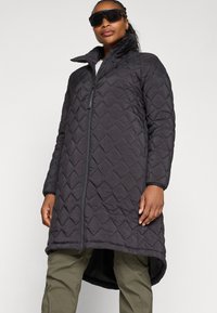 Quilted black long coat with high collar and zipper closure. Features a diamond pattern, elastic cuffs, and a curved hem for added length.
