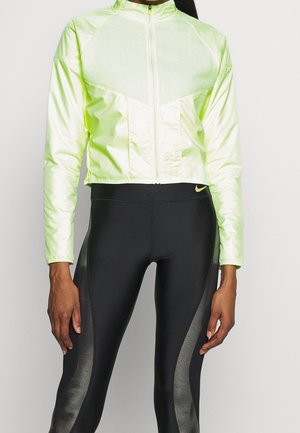 Kobieta w jasnoszarozielonej kurtce zapinanej na zamek oraz czarnych legginsach z szarymi panelami i logo Nike na pasie.