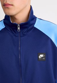 Casaco com fecho da Nike em azul-marinho com realces em azul-claro nos ombros. Apresenta um colarinho alto, tecido suave e um emblema quadrado no peito.