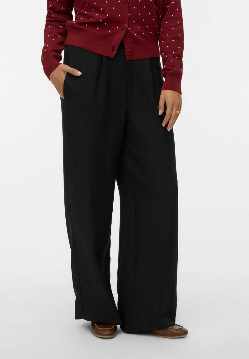 Personne portant un pantalon large noir ample avec les mains partiellement dans les poches, associé à des chaussures brunes et un cardigan rouge à pois blancs.