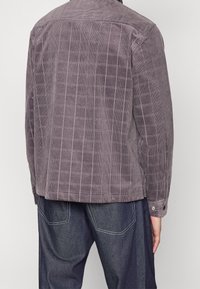 Chaqueta de pana gris con un patrón de cuadros, que presenta un cuello con botones, aberturas laterales y puños abotonados, combinada con pantalones de mezclilla oscuros.