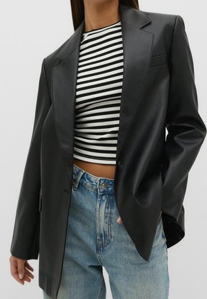 Femme portant un blazer en cuir noir, un crop top rayé noir et blanc, et un jean en denim bleu, se tenant avec une main dans la poche.