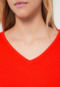 Κόκκινο πλεκτό πουλόβερ με ντεκολτέ σε σχήμα V, με απαλό υλικό και εφαρμοστή γραμμή. Δεν φαίνονται επιπλέον μεταλλικά στοιχεία ή σχέδια.