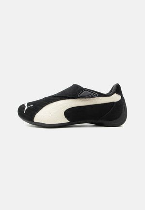 Zapatilla Puma blanca y negra tipo slip-on con detalle de franja blanca curvada y suela de goma redondeada sobre un fondo claro liso.