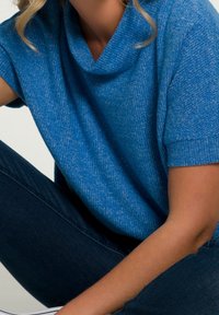 Maglione azzurro a collo alto realizzato in tessuto testurizzato, con maniche corte. Indossato con jeans scuri, che mettono in mostra un fit casual e rilassato.