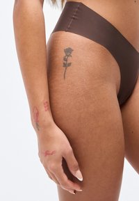 Καφέ μαγιό με ψηλή μέση και λεία υφή, με ψηλή κοπή. Δύο τατουάζ ορατά στο χέρι: ένα τριαντάφυλλο και μια λέξη.