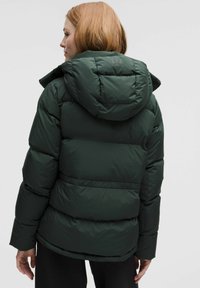 Roheline puffer-jope kapuutsiga, tikitud sektsioonidega ja brändi logoga. Materjal näib olema sile ja soojustatud.