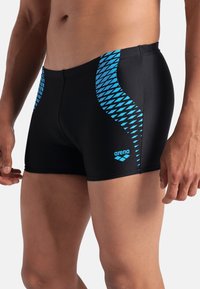 Pantaloni da nuoto neri con dettagli geometrico blu, realizzati in materiale elasticizzato. Presentano un taglio aderente con un logo vicino all'orlo.