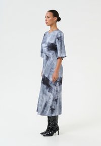 Abito lungo grigio con un motivo tie-dye e dettagli neri, maniche ampie e una silhouette snella, abbinato a stivali neri lucidi al ginocchio.