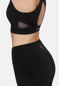 Black sports bra with a cut-out mesh design and logo accent. Paired with black high-waisted leggings featuring stitching details and logo.