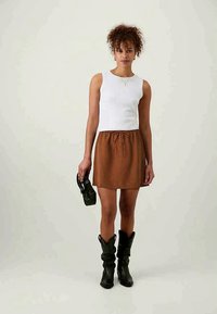 Haut sans manches blanc associé à une jupe à cordon marron. Modèle portant des bottes noires amples, tenant un sac à main noir à découpe.