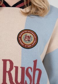 Pull en tricot avec rayures verticales beige et bleu clair, lettrage rouge et écusson rond portant l'inscription "Prohibited Motorsport Company Berlin Germany".