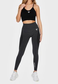 Sostenedor deportivo negro con tirantes finos, combinado con leggings negros de cintura alta que presentan un patrón texturizado y el texto "SQUATPROOF" en blanco.