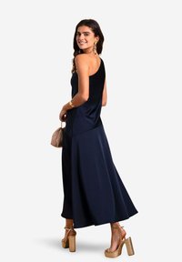 Robe en satin marine avec une conception à une épaule, jupe fluide et texture lisse. Associée à des talons dorés épais et à un petit sac à main beige.